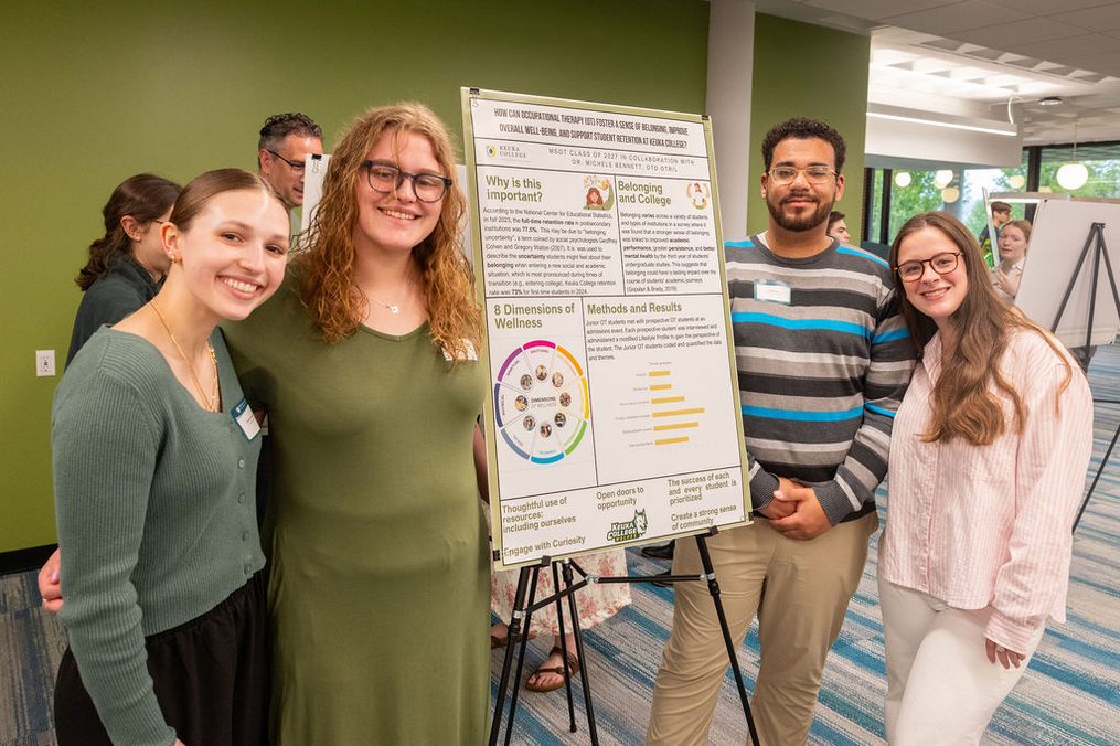 Four college students stand smiling on either side of a research poster displayed on an easel in a campus hallway. The poster discusses occupational therapy, student belonging, and wellness, with charts and text sections. The students wear name tags and casual attire, and other attendees are visible in the background of the indoor event space.