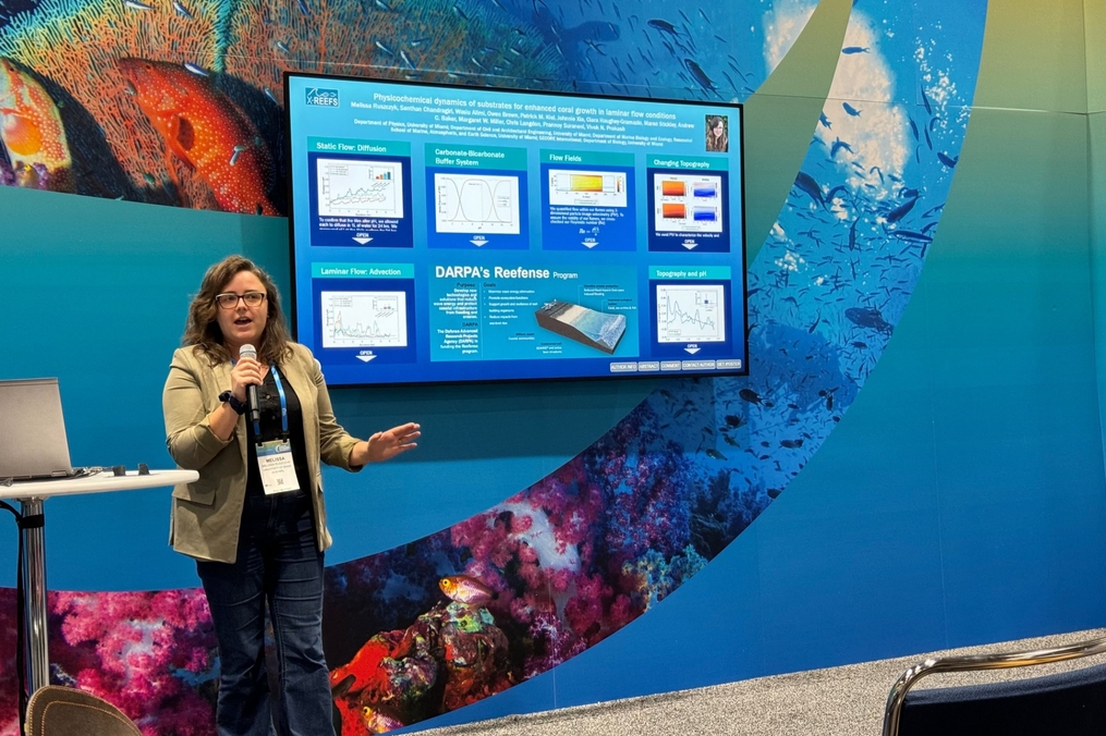 A person stands at the front of a conference presentation area, speaking into a microphone and gesturing toward a large digital poster displayed on a screen behind them. The presentation appears to focus on coral reef research, with charts, graphs, and diagrams visible on the screen. The backdrop features colorful underwater imagery, including coral reefs and fish, creating an ocean-themed setting. A laptop sits on a nearby table, and audience seating is visible in the foreground.