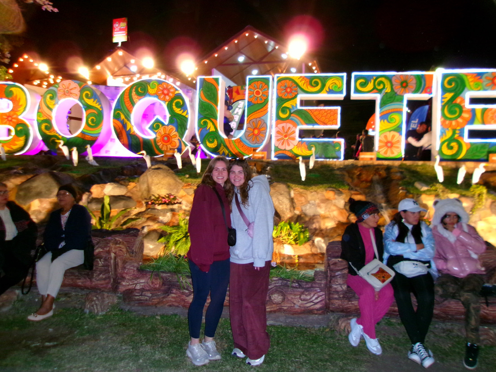 Two people posing in front of colorful illuminated “Boquete” letters at night, with festival lights and visitors in the background.